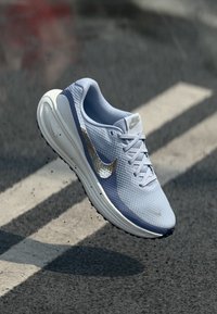 Svijetloplavi Nike tenisica za trčanje s metalik swooshom lebdi iznad asfaltnog puta s bijelim trakama za prelazak.