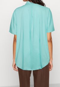 Camicia a maniche corte color acquamarina con vestibilità rilassata, dettaglio della piega sul retro, colletto ordinato e orlo arrotondato, realizzata in un tessuto leggero e liscio.