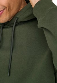 Mørkegrøn sweatshirt med et struktureret stof, der har en foret hætte og matchende snore. Nærbillede af halsudskæring og skulderområde.