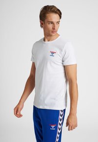 Mand iført hvid kortærmet skjorte og blå træningsbukser med røde og hvide Hummel-logoer, stående og kigger mod højre.