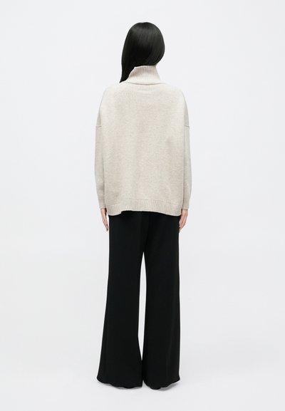 Γυναίκα με μακριά σκούρα μαλλιά που φοράει πουλόβερ ζιβάγκο μπεζ και μαύρο παντελόνι με φαρδιά μπατζάκια, στέκεται γυρισμένη με την πλάτη σε απλό φόντο.