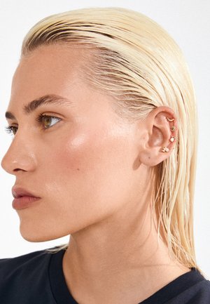 Plava osoba s zalizanom mokrom kosom koja nosi pet malih zlatnih naušnica u obliku polumjeseca, sunca, školjke i zvijezda.