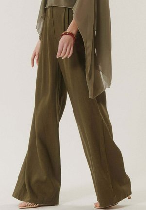 Femme portant un pantalon large olive, un haut à manches cape transparentes, des talons dorés à brides, et des bracelets rouges et dorés au poignet gauche.