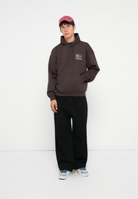 Mørkegrå hættebluse med frontlomme og logo, parret med løse sorte bukser og lyseblå sneakers. Accessoriseret med en lyserød kasket.