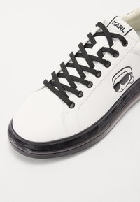 Biały sneaker z czarnymi sznurowadłami i wykończeniem wokół dziurek, z przezroczystą czarną podeszwą i graficznym logo po boku.