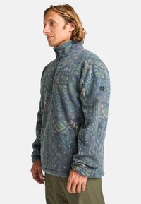 Fleece jacket in blue and green with geometric patterns, high collar, zip front, and side pockets. Soft texture and relaxed fit.