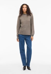 Γυναίκα με μακριά σγουρά μαλλιά, φορώντας έναν ταμπά πουλόβερ, μπλε τζιν με ίσια γραμμή και μαύρες γόβες, στέκεται μπροστά σε λευκό φόντο.