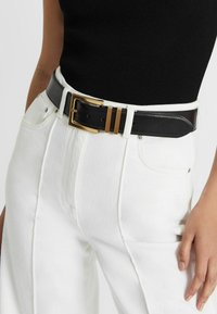 Schwarzer Ledergürtel mit einer goldenen Metallschnalle, die drei horizontale Streifen aufweist, getragen über weißen Denim-Hosen.