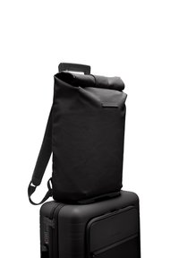 Sac à dos noir à rabat, fabriqué en matériau texturé, doté de bretelles réglables et d'un design minimaliste. Posé sur une valise noire.