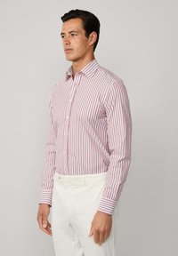 Bílá košile s červenými svislými pruhy, s klasickým límečkem a dlouhými rukávy, v kombinaci s bílými kalhotami.