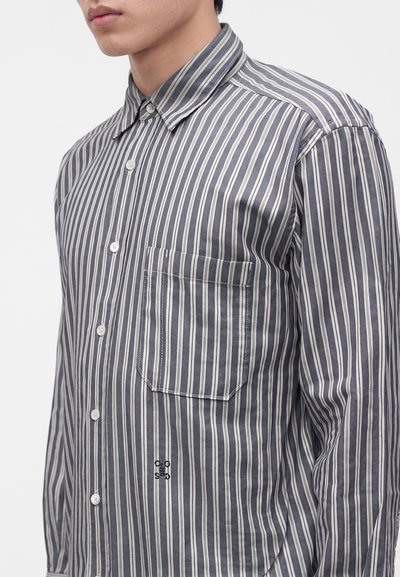 Homme portant une chemise à rayures verticales grises et blanches avec une poche poitrine et un petit logo brodé.