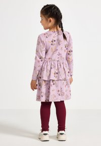 Młoda dziewczynka z warkoczami, ubrana w lawendową sukienkę w kwiaty, bordowe legginsy prążkowane i białe sneakersy, stoi z plecami do widza.