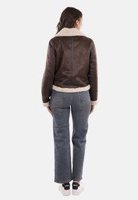 Mujer de espaldas, vistiendo una chaqueta de piel de oveja marrón, jeans rectos grises y zapatillas blancas con detalle negro en el talón.