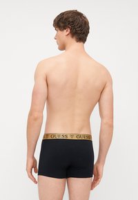 Melnas apakšbikses ar zeltītu "GUESS" zīmolu jostu, ar pieguļošu dizainu un mīkstu auduma tekstūru. Skatīts no aizmugures.