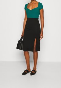 Kobieta ubrana w turkusowy top z kwadratowym dekoltem i czarną spódnicę z wysokim stanem oraz przednim rozcięciem, trzyma czarną torebkę i nosi czarne spiczaste baleriny.