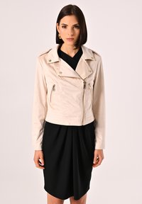 Giacca di pelle cropped beige chiaro con tasche con zip, colletto a revers e dettagli metallici. Indossata sopra un vestito nero.