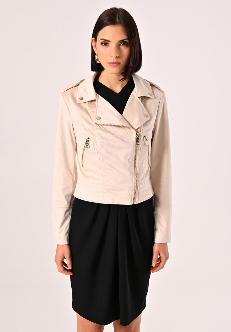 Giacca di pelle cropped beige chiaro con tasche con zip, colletto a revers e dettagli metallici. Indossata sopra un vestito nero.