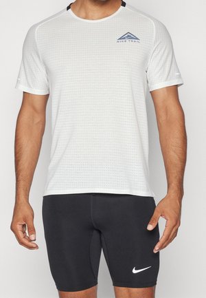 Mężczyzna w białej, krótkiej koszulce Nike Trail oraz czarnych sportowych szortach Nike, stojący z rękami swobodnie opuszczonymi wzdłuż ciała.
