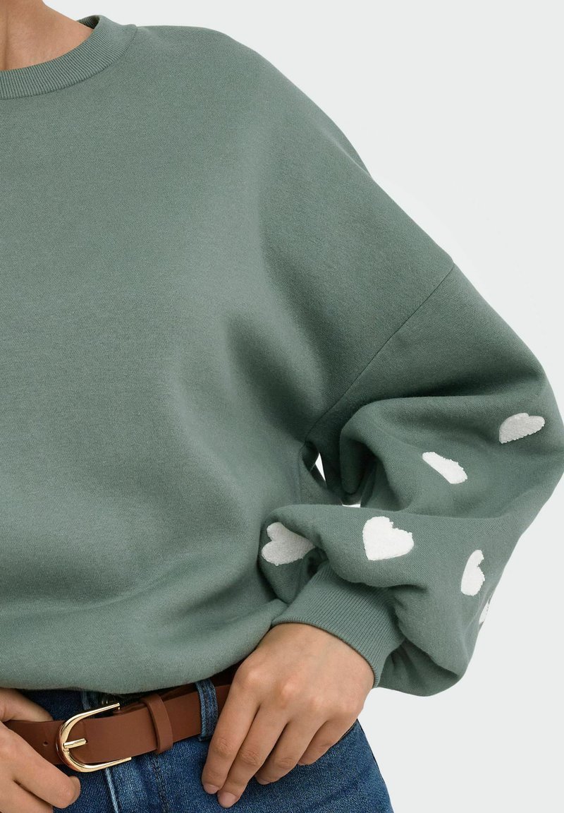 Personne portant un sweat-shirt vert avec des motifs de cœurs blancs sur les manches, rentré dans un jean bleu avec une ceinture marron.