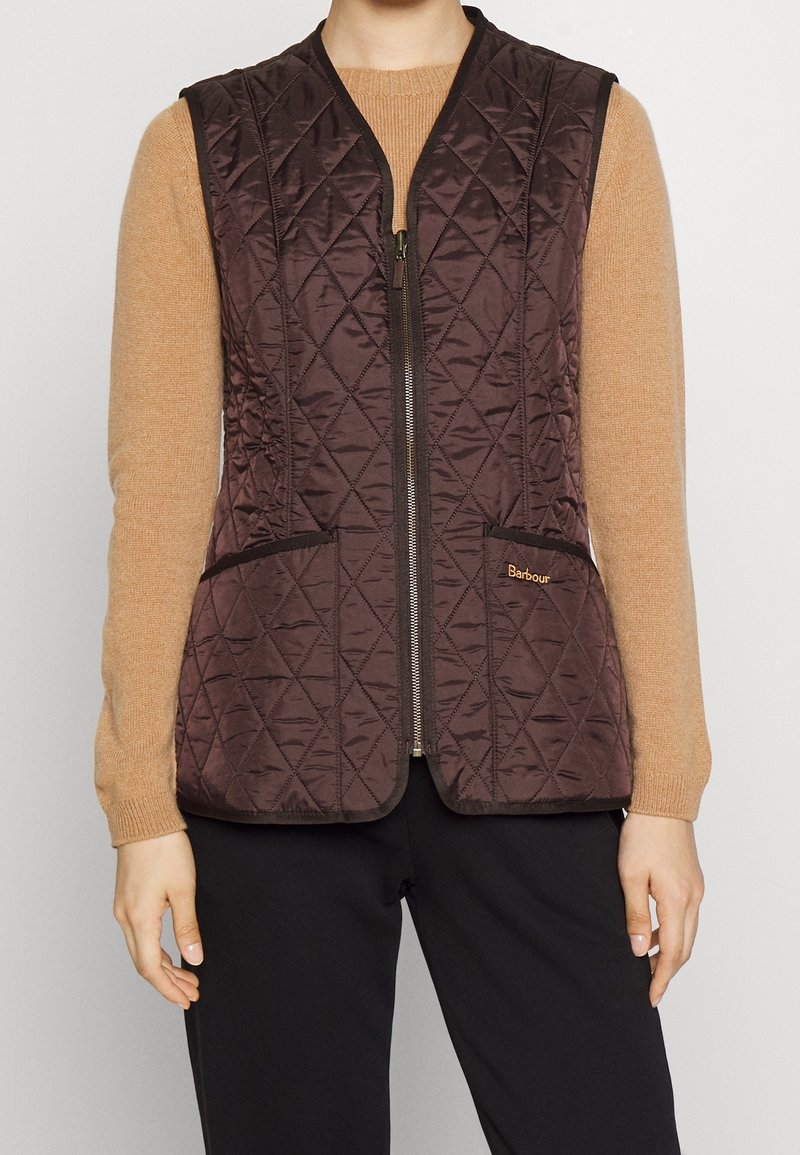 Femme portant une veste sans manches matelassée marron sur un pull beige à manches longues et un pantalon noir, debout les bras détendus le long du corps.
