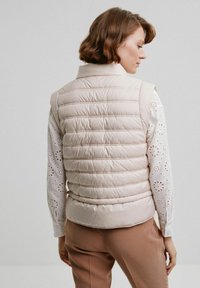 Beżowa pikowana kamizelka z wysokim kołnierzem, z poziomymi kanałami i gładką teksturą, noszona na białej koszuli w wzór oczek.