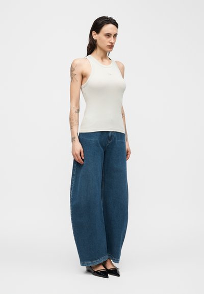 Γυναίκα με τατουάζ που φοράει λευκό αμάνικο ριμπ τοπ, φαρδιά μπλε τζιν παντελόνια και μαύρες μυτερές φλατ, όρθια μπροστά σε απλό φόντο.