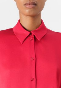 Blouse rose vif avec un col classique replié, cinq boutons sur le devant et une texture lisse, semblable à du satin.