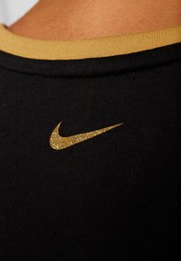 Maglietta nera in tessuto con un logo Nike in glitter dorato sul retro. Presenta un colletto a costine dorato a contrasto sullo scollo.