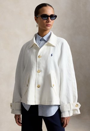 Γυναίκα με υπερμεγέθη λευκό σακάκι με κουμπιά πάνω από μπλε ριγωτό πουκάμισο, μαύρο παντελόνι και μαύρα γυαλιά ηλίου στυλ cat-eye, στέκεται μπροστά σε ουδέτερο φόντο.