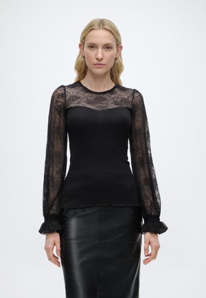 Blondynka w czarnej bluzce z koronkowymi, przezroczystymi, kwiatowymi, długimi rękawami oraz dopasowanej czarnej skórzanej spódnicy, stojąca na białym tle.