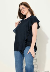 Granatowa bluzka z falbaniastymi rękawami i panelami z przodu; zestawiona z jasnoniebieskimi dżinsami. Modelka nosi okulary, prezentując swobodny strój.