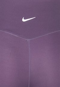 Fioletowe legginsy sportowe wykonane z rozciągliwego materiału o gładkiej teksturze, z białym logo Nike na pasie i szwami flatlock.