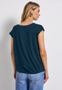 T-shirt bleu-vert à coupe décontractée, à manches courtes et col arrondi, associé à un jean en denim bleu clair, présenté sous un angle latéral-arrière.