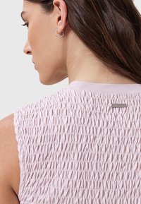 Ljust rosa strukturerad ärmlös topp med smockdetalj bak och en liten metallisk varumärkesetikett. Enkelt runt halslinning.