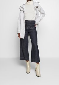 Světle šedá nepromokavá bunda přes krémový rolák, kombinovaná s tmavě modrými širokými džínami a krémovými kotníkovými botami se vzorem hadí kůže.