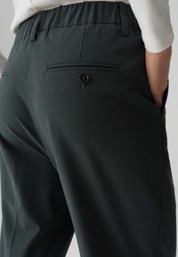 Dunkelgrüne Hose mit weicher Textur, ausgestattet mit einer Gesäßtasche mit Knopf und einem elastischen Bund für eine verstellbare Passform.