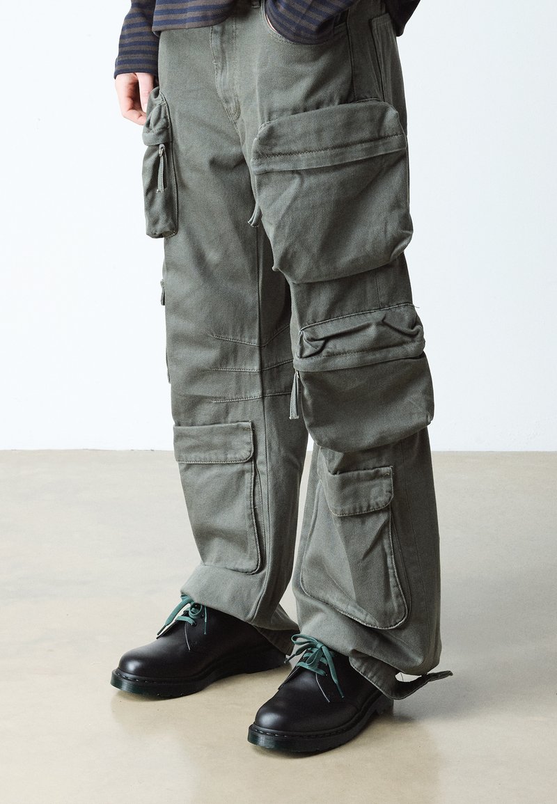Personne portant un pantalon cargo vert olive avec plusieurs grandes poches et des chaussures noires à lacets avec des lacets verts sur un sol lisse.