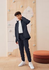 Navy blauwe zip-up jas en bijpassende sweatpants gemaakt van zachte stof, gedragen met een wit t-shirt en witte sneakers. Achtergrond van een klimwand.