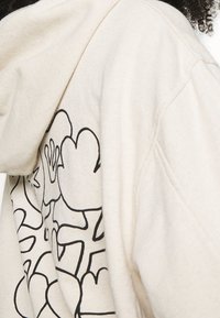 Sweat à capuche beige avec une texture douce, présentant un motif graphique floral noir sur le côté. Le design comprend des formes abstraites et des contours.