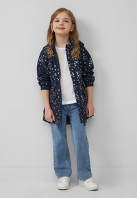 Granatowa kurtka przeciwdeszczowa w kwiaty z elastycznymi mankietami i zamkiem, noszona na białej koszuli i jasnoniebieskich dżinsach, w połączeniu z białymi sneakersami.