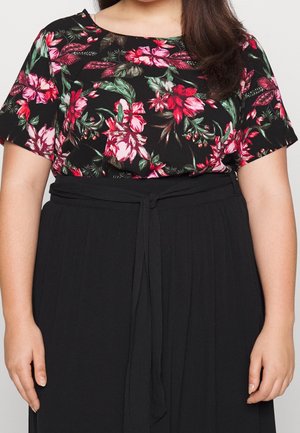 Kobieta ubrana w czarny top w kwiaty z krótkim rękawem, włożony do wysokich czarnych spodni z paskiem wiązanym w talii.