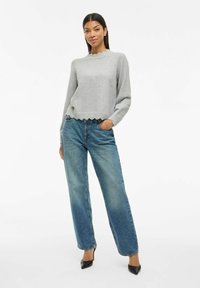 Kvinna står med handen i fickan iförd en grå tröja med scallopkant, blå raka jeans och svarta spetsiga klackar mot vit bakgrund.