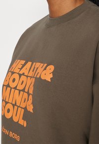 Gros plan sur une chemise marron avec un texte orange vif affichant « Health & Body & Mind & Soul » et un petit logo « Björn Borg » en dessous.