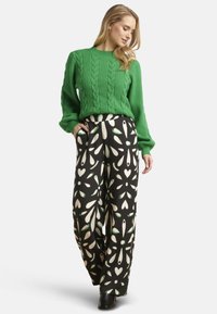 Green cable-knit sweater paired with black wide-leg trousers featuring a white and green leaf pattern. Model wears black ankle boots.