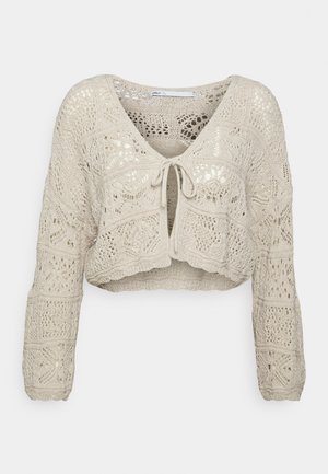 Cárdigan recortado en beige claro con intrincados patrones de punto. Presenta un escote en V, mangas largas y un cierre con lazo en la parte delantera.