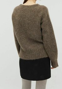 Personne portant un pull brun doux, une jupe noire et des collants côtelés gris clair, vue de dos sur un fond uni.