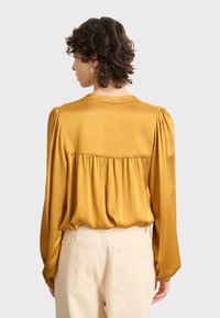 Persona con cabello corto y rizado, vistiendo una blusa dorada de satén de manga larga y pantalones de cintura alta color crema, de espaldas sobre un fondo sencillo.