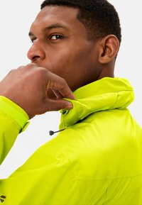 Neon groene jas met hoge kraag, gestructureerde stof, verstelbare capuchon en contrasterende zwarte hardware. Close-up van de hand van een persoon die de kraag vasthoudt.