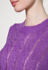 Pull-over en tricot violet vif avec un motif texturé et ajouré. Dispose d'un col ras du cou côtelé et d'un design complexe en torsades.