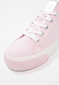 Różowy tenisówka na płótnie z białą gumową podeszwą, zaokrąglonym noskiem i białymi sznurówkami. Wyróżnia się srebrnym logo i teksturowanymi bocznymi akcentami.